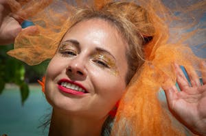 Close-up of a woman smiling with glitter makeup and an orange tulle veil, enjoying a sunny day.