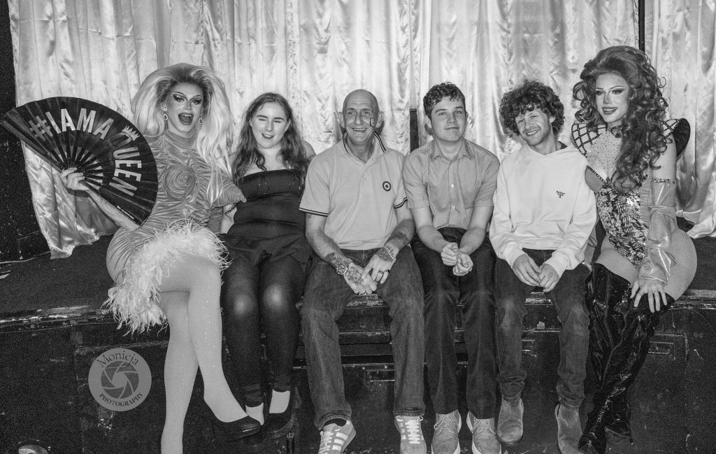 Two drag queens posing with four audience members on stage during the Penicuik charity night, a joyful group photo celebrating inclusivity and community.