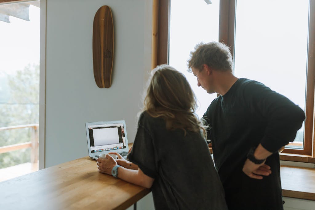 A man and woman stand at a kitchen counter, working on a laptop near a sunny window.