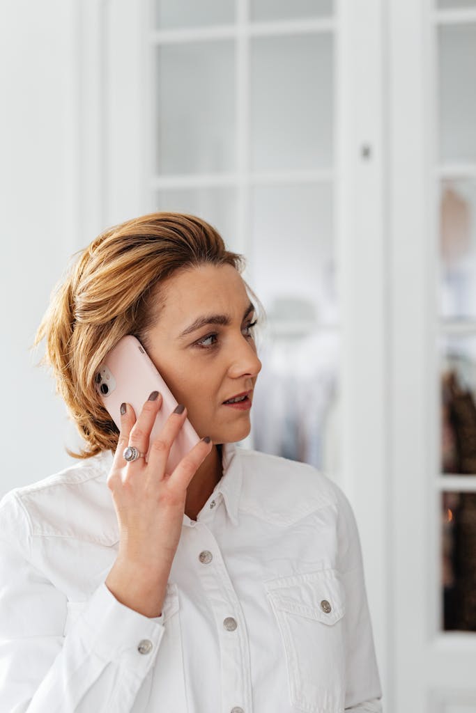 Adult woman in a white shirt engaged in a phone call on a smartphone indoors.