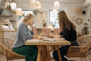 Two women collaborating and writing in a cozy cafe setting over coffee and pastries.
