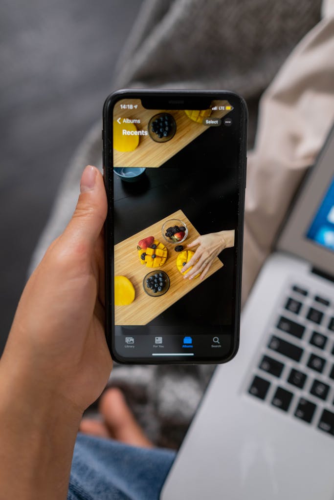 Photo by Ron Lach Close-up of a hand holding a smartphone showing a vibrant fruit platter on a laptop background.