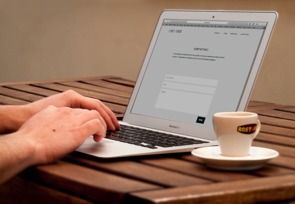 Close-up of hands typing on a laptop next to a cup of coffee on a wooden desk.